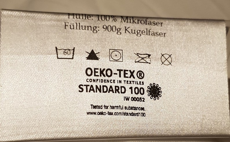 May include: A silver-coloured fabric care label featuring the text H&uuml;lle: 100% Mikrofaser, F&uuml;llung: 900g Kugelfaser, and OEKO-TEX Standard 100. The label includes five laundry care symbols, the OEKO-TEX logo, and the website www.oeko-tex.com/standard100.