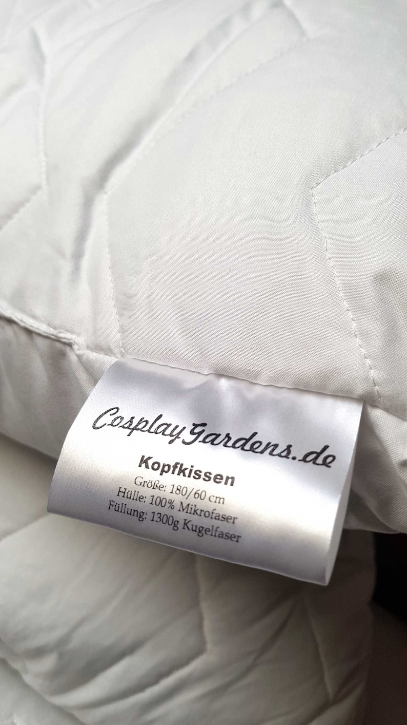 May include: A white rectangular pillow with a quilted surface pattern. A white fabric tag is attached, featuring the text CosplayGardens.de, Kopfkissen, Gr&ouml;&szlig;e: 180/60 cm, H&uuml;lle: 100% Mikrofaser, and F&uuml;llung: 1300g Kugelfaser in black printed lettering.