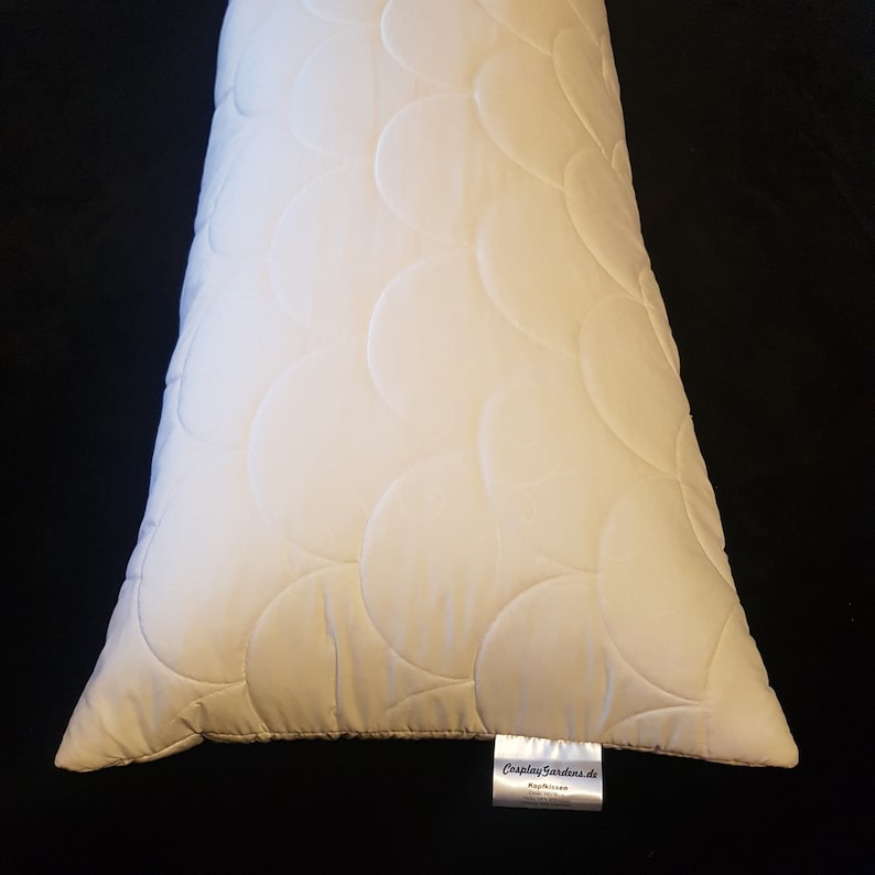 May include: A rectangular off-white pillow featuring a quilted pattern of overlapping circles. A small white fabric tag is attached to the bottom edge, displaying the text CosplayGardens.de, Kopfkissen, 150x50cm, 100% Polyester.