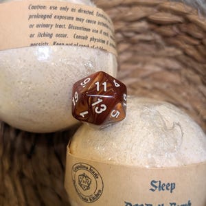 May include: Two round, off-white bath bombs wrapped in clear plastic with tan paper bands. One band features the text "Grandma Jean's Kitchen" and "Sleep D20 Bath Bomb". A brown and gold marbled twenty-sided die sits between the two bath bombs.