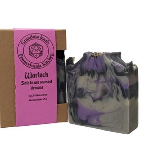 May include: A bar of handmade soap with purple and gray swirls, next to a brown box with a pink label. The label reads "Grandma Jean's Pennsylvania Kitchen Warlock Safe to use on most demons".