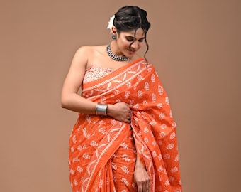 May include: A woman wearing an orange and white cotton saree with a floral print. The saree is draped over her shoulders and waist, and she is standing with her head slightly bowed.
