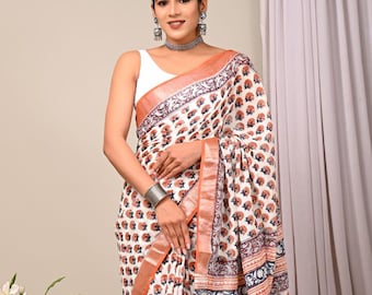 May include: White saree with a repeating floral pattern in orange and blue. The saree has a rust-colored border and a detailed patterned edge. The garment is draped, showcasing the design and texture of the fabric. The saree is a traditional garment.