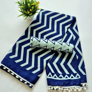 May include: A blue and white cotton saree with a zig-zag pattern and white pom-poms along the border. The saree is folded in half and has a matching blue and white fabric piece on top.