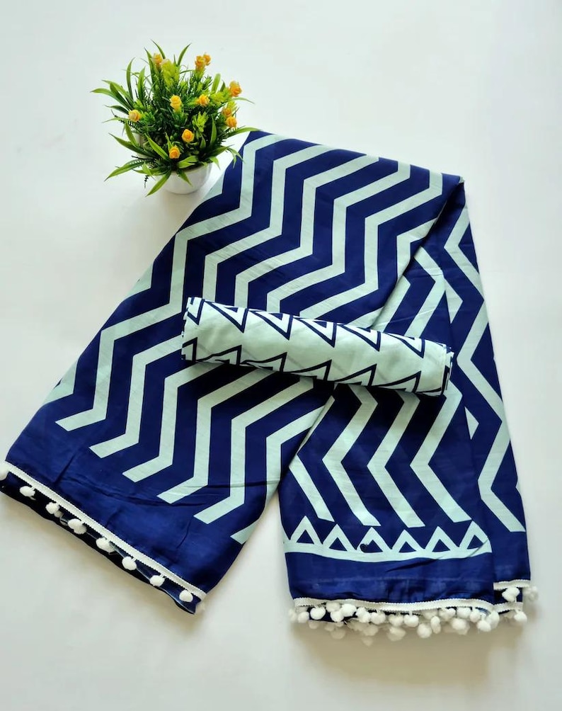 May include: A blue and white cotton saree with a zig-zag pattern and white pom-poms along the border. The saree is folded in half and has a matching blue and white fabric piece on top.