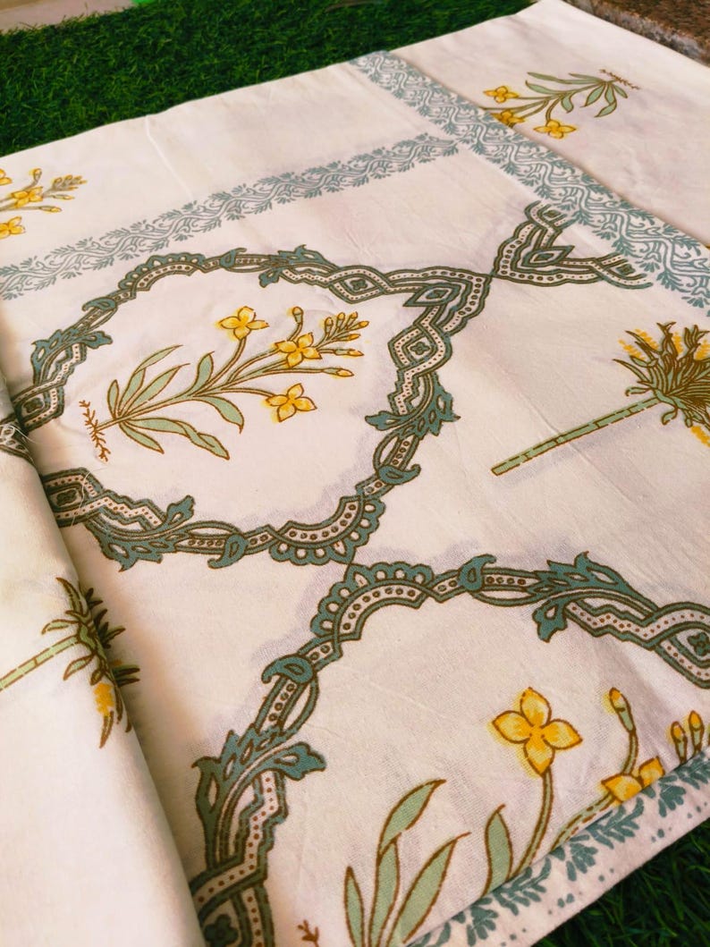 Puede incluir: Tela blanca con un estampado floral repetido en amarillo, verde y verde azulado. El dise&ntilde;o presenta flores y hojas estilizadas dentro de marcos geom&eacute;tricos ornamentados. Los bordes est&aacute;n bordeados con un delicado patr&oacute;n verde azulado.