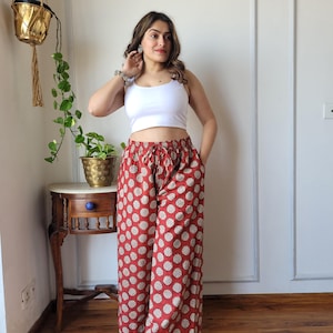 Puede incluir: Una mujer con un top blanco corto y pantalones palazzo rojos con estampado floral blanco. Está de pie en una habitación con suelo de madera y una mesa auxiliar.