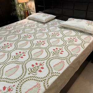 May include: A white bedsheet featuring a repeating pattern of olive green leaves and red floral clusters, enclosed in a circular frame. Two matching pillowcases are visible. The bedsheet has a tan border.