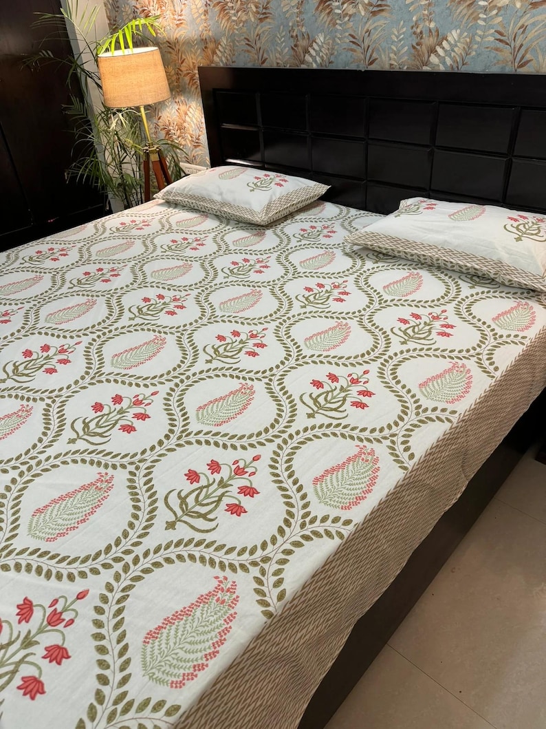 May include: A white bedsheet featuring a repeating pattern of olive green leaves and red floral clusters, enclosed in a circular frame. Two matching pillowcases are visible. The bedsheet has a tan border.