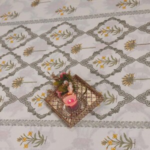 Puede incluir: Mantel blanco con un patr&oacute;n repetido de ramitas florales amarillas y bordes geom&eacute;tricos grises. Una peque&ntilde;a bandeja dorada contiene una vela rosa encendida y un arreglo floral, agregando un toque de color.