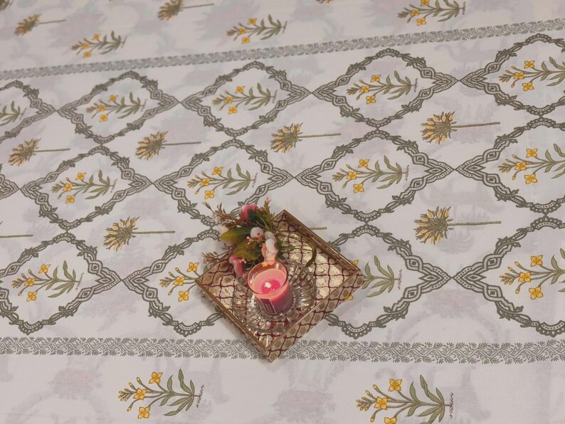 Puede incluir: Mantel blanco con un patr&oacute;n repetido de ramitas florales amarillas y bordes geom&eacute;tricos grises. Una peque&ntilde;a bandeja dorada contiene una vela rosa encendida y un arreglo floral, agregando un toque de color.