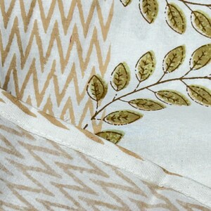 May include: Close-up of a fabric with two distinct patterns. One section features a repeating zigzag design in tan and white. The other displays a white background with brown branches and olive-green leaves. The fabric is likely used for home decor.