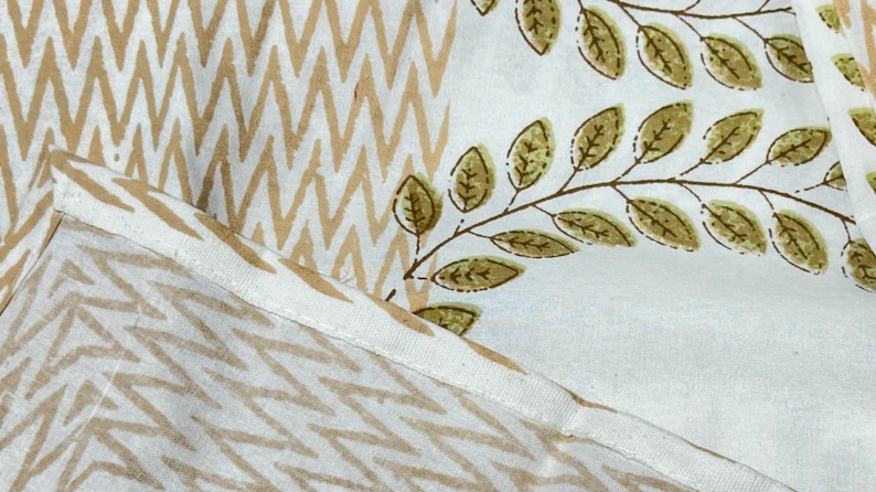 May include: Close-up of a fabric with two distinct patterns. One section features a repeating zigzag design in tan and white. The other displays a white background with brown branches and olive-green leaves. The fabric is likely used for home decor.