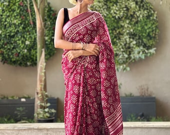 May include: A maroon linen saree with a white floral print. The saree has a silver border and is draped on a woman.