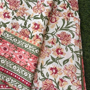 Può includere: Tessuto bianco con un motivo floreale nei toni del rosso, pesca e giallo, con foglie e viti verdi. È visibile un bordo con strisce rosa e verdi e motivi geometrici. Il tessuto è piegato e impilato.