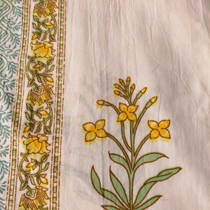 Puede incluir: Primer plano de una tela blanca con un estampado floral. El dise&ntilde;o presenta flores amarillas con hojas y tallos verdes. Un borde con flores amarillas y hojas verdes est&aacute; en el lado izquierdo. La tela tiene un aspecto suave y texturizado.