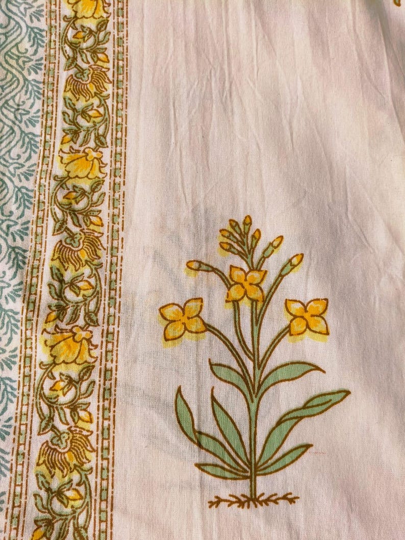Puede incluir: Primer plano de una tela blanca con un estampado floral. El dise&ntilde;o presenta flores amarillas con hojas y tallos verdes. Un borde con flores amarillas y hojas verdes est&aacute; en el lado izquierdo. La tela tiene un aspecto suave y texturizado.