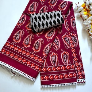 May include: A burgundy textile with a paisley pattern in white, orange, and red. A black and white diamond-patterned accessory rests on top. The edges are trimmed with white pom-poms. This textile is suitable for clothing or home decor.