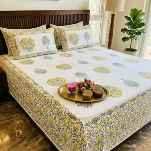May include: White bed linens with a yellow and gray floral pattern. The set includes a fitted sheet, pillowcases, and a decorative tray with a candle and flowers. The bed is made up with a dark wood headboard.