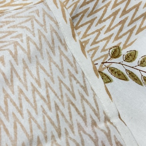 May include: Close-up of white fabric with a repeating zigzag pattern in light brown. A section of the fabric features a branch with green leaves. The fabric is likely used for home decor or textiles.
