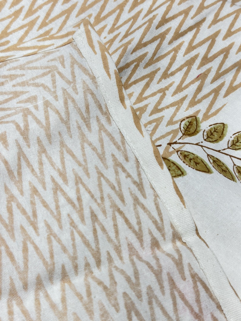 May include: Close-up of white fabric with a repeating zigzag pattern in light brown. A section of the fabric features a branch with green leaves. The fabric is likely used for home decor or textiles.