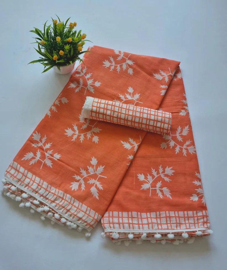 May include: Orange cotton saree with white leaf print and white checkered border. The saree has a white pom-pom trim.