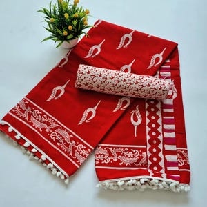 May include: Red and white cotton saree with a white floral print and a white pom-pom trim. The saree is folded and displayed on a white surface.