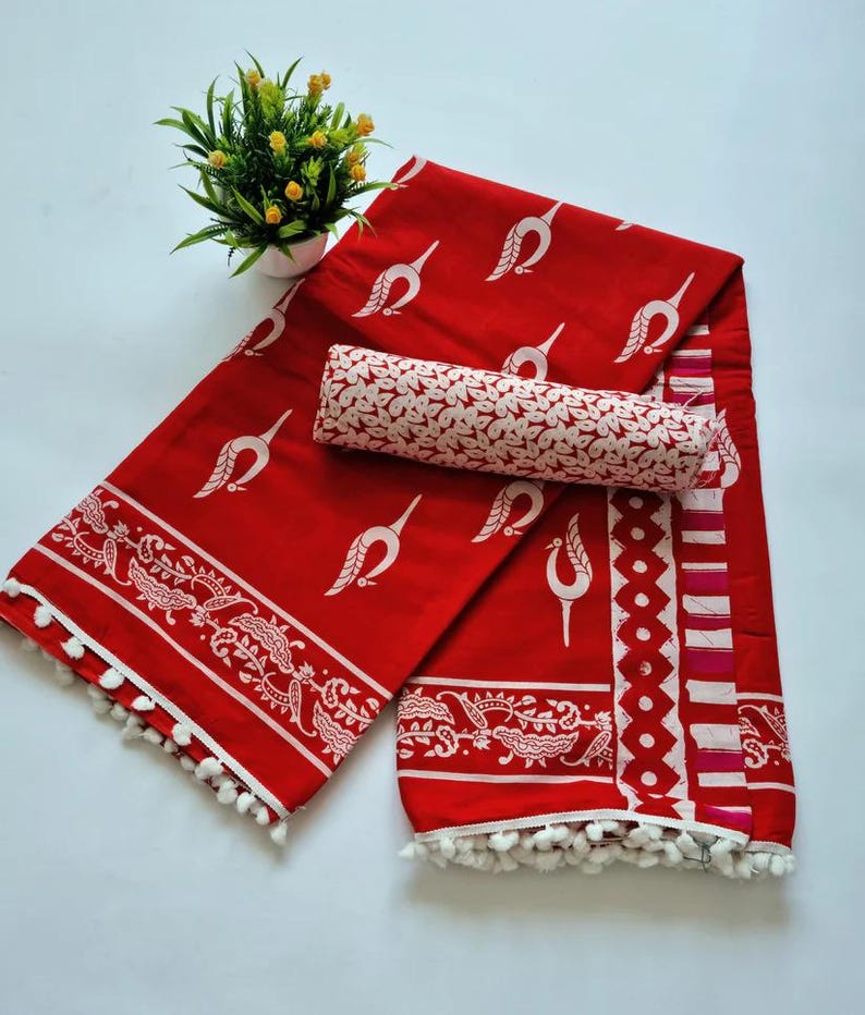 May include: Red and white cotton saree with a white floral print and a white pom-pom trim. The saree is folded and displayed on a white surface.