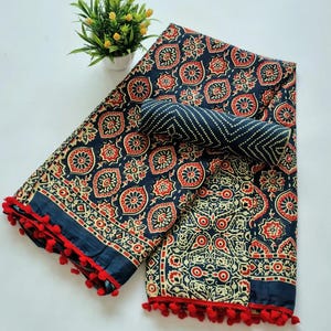May include: A blue and white cotton saree with a red and white floral pattern. The saree has a pom-pom trim and is folded in half.