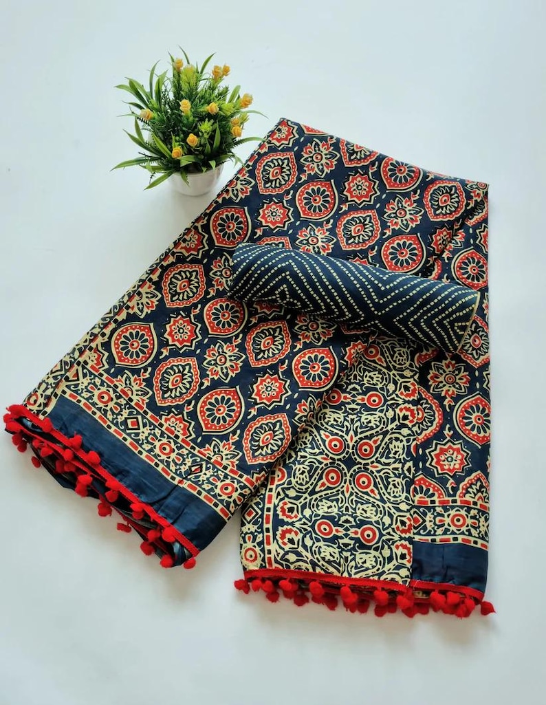 May include: A blue and white cotton saree with a red and white floral pattern. The saree has a pom-pom trim and is folded in half.