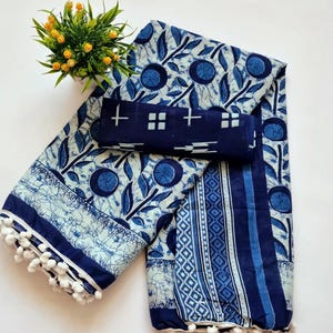 May include: A blue and white cotton saree with a floral print and a white pom-pom trim. The saree is folded in half and has a matching blouse piece.