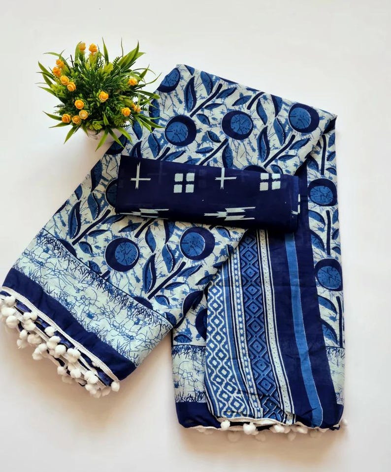 May include: A blue and white cotton saree with a floral print and a white pom-pom trim. The saree is folded in half and has a matching blouse piece.
