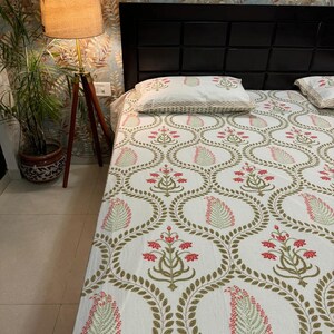 May include: A white bedspread and pillowcases featuring a repeating pattern of red floral and green leaf designs, enclosed in gold oval shapes. The bed is set against a black headboard and floral wallpaper.