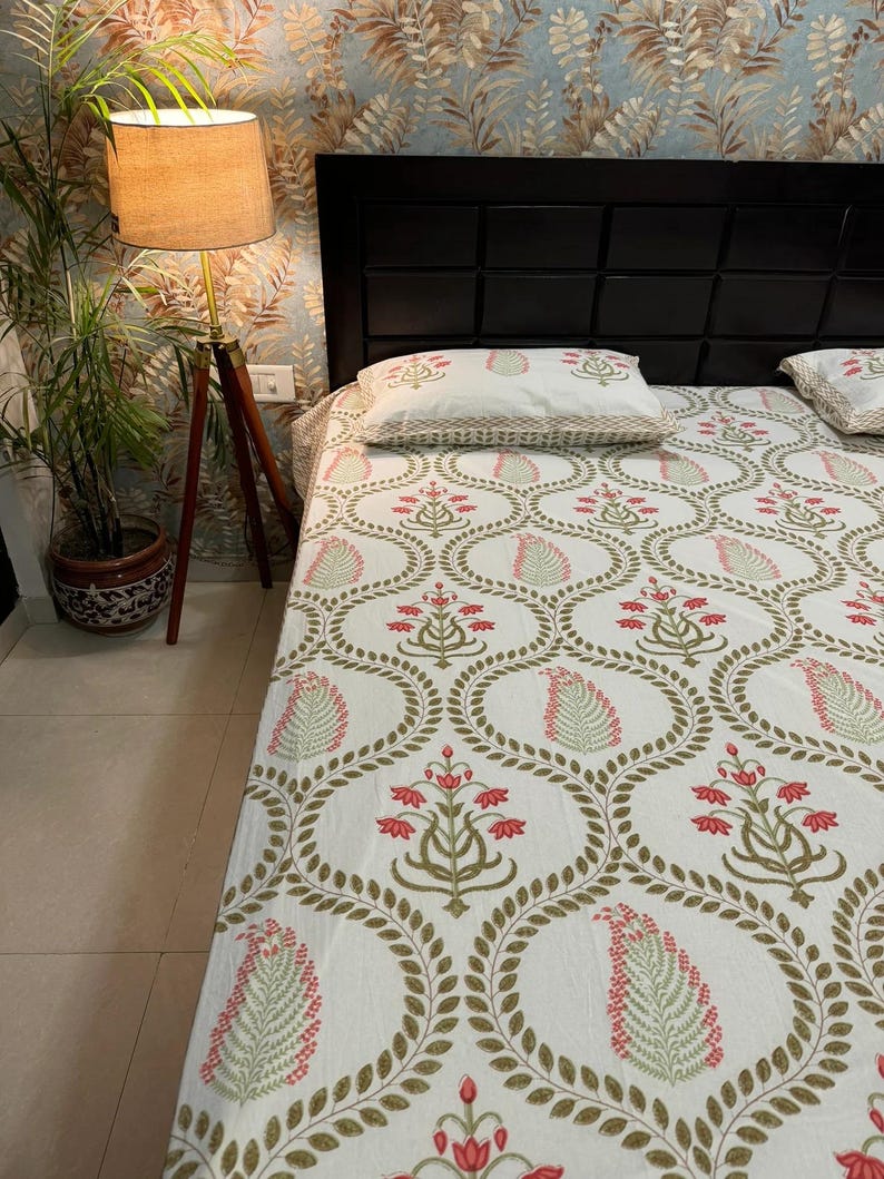 May include: A white bedspread and pillowcases featuring a repeating pattern of red floral and green leaf designs, enclosed in gold oval shapes. The bed is set against a black headboard and floral wallpaper.