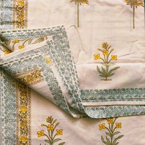 Puede incluir: Tela de color crema con un estampado floral repetido en amarillo y verde. El dise&ntilde;o incluye flores estilizadas y motivos de &aacute;rboles. Los bordes est&aacute;n bordeados con un patr&oacute;n detallado en verde y amarillo.
