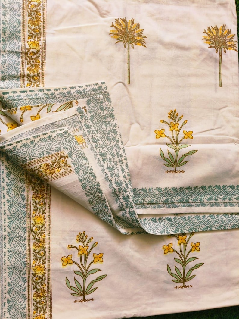 Puede incluir: Tela de color crema con un estampado floral repetido en amarillo y verde. El dise&ntilde;o incluye flores estilizadas y motivos de &aacute;rboles. Los bordes est&aacute;n bordeados con un patr&oacute;n detallado en verde y amarillo.
