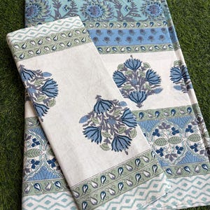 Peut inclure: Un textile plié présentant un motif floral imprimé au bloc dans des tons de bleu, vert sauge et blanc. Le design comprend des motifs répétitifs de fleurs stylisées, des bordures géométriques en chevron et des motifs de feuilles complexes.