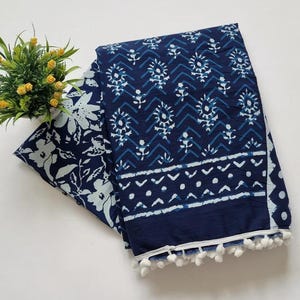 May include: A blue and white cotton fabric with a floral pattern on one side and a geometric pattern on the other. The fabric has a white pom-pom trim.