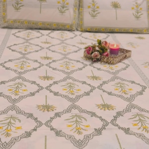 Puede incluir: Juego de ropa de cama con estampado floral blanco, que incluye dos fundas de almohada. La colcha presenta un diseño repetido de flores amarillas y verdes dentro de un borde geométrico gris. Una pequeña bandeja con flores y una vela está sobre la cama.