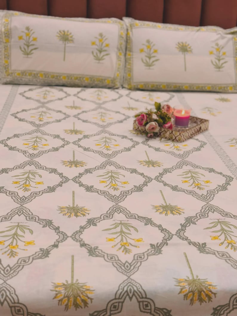 Puede incluir: Juego de cama blanco con un patr&oacute;n repetido de motivos florales amarillos y verdes dentro de formas de diamante grises. Las fundas de almohada tienen un borde amarillo. Una peque&ntilde;a bandeja con flores y una vela est&aacute; sobre la cama.