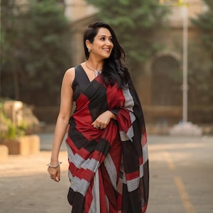 May include: A woman wearing a black, red, and grey patterned saree. The saree has a geometric design with bold stripes of black, red, and grey. The woman is smiling and wearing jewellery.