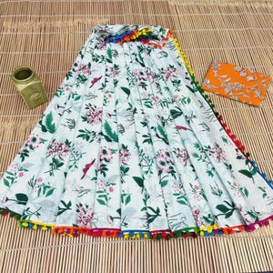 May include: A white fabric with a floral pattern of pink flowers and green leaves. The fabric is trimmed with a colorful pom-pom border. An orange patterned pouch and a green jar are also visible.