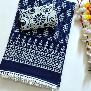 May include: A folded navy blue fabric featuring a white floral and geometric block print pattern. The fabric includes a distinct border with a repeating motif and is finished along the bottom edge with a white decorative fringe trim.