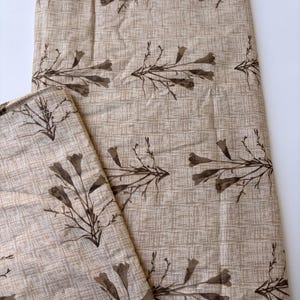 May include: A piece of light tan fabric featuring a repeating dark brown botanical print of small, delicate flowers on thin stems. The textile has a subtle, cross-hatched woven texture throughout, creating a grid-like pattern across the surface.