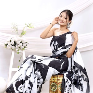 Peut inclure: Un sari imprimé en noir et blanc avec des visages répétés. Le sari est drapé sur une personne assise sur un tabouret décoratif. La personne porte un haut à une épaule fait du même tissu. Un bouquet de fleurs blanches est à proximité.