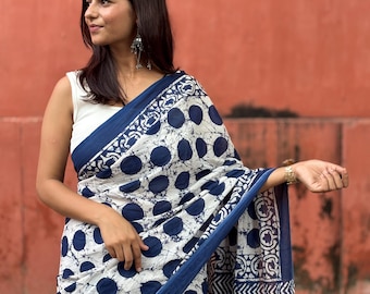 Könnte beinhalten: Ein weißer und marineblauer Sari mit großem Polka-Dot-Muster. Der Sari hat einen marineblauen Rand mit geometrischen und floralen Designs. Die Frau trägt ein weißes Oberteil und silberne Ohrringe.