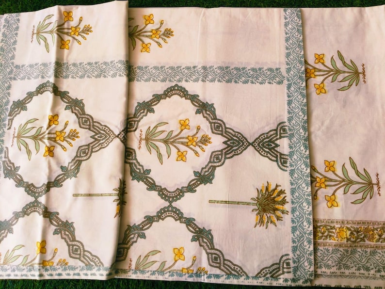 Puede incluir: Tela blanca con un patr&oacute;n repetitivo de flores amarillas y hojas verdes. El dise&ntilde;o est&aacute; enmarcado por un borde de elementos decorativos verdes y azules. La tela est&aacute; doblada, mostrando la repetici&oacute;n del patr&oacute;n.