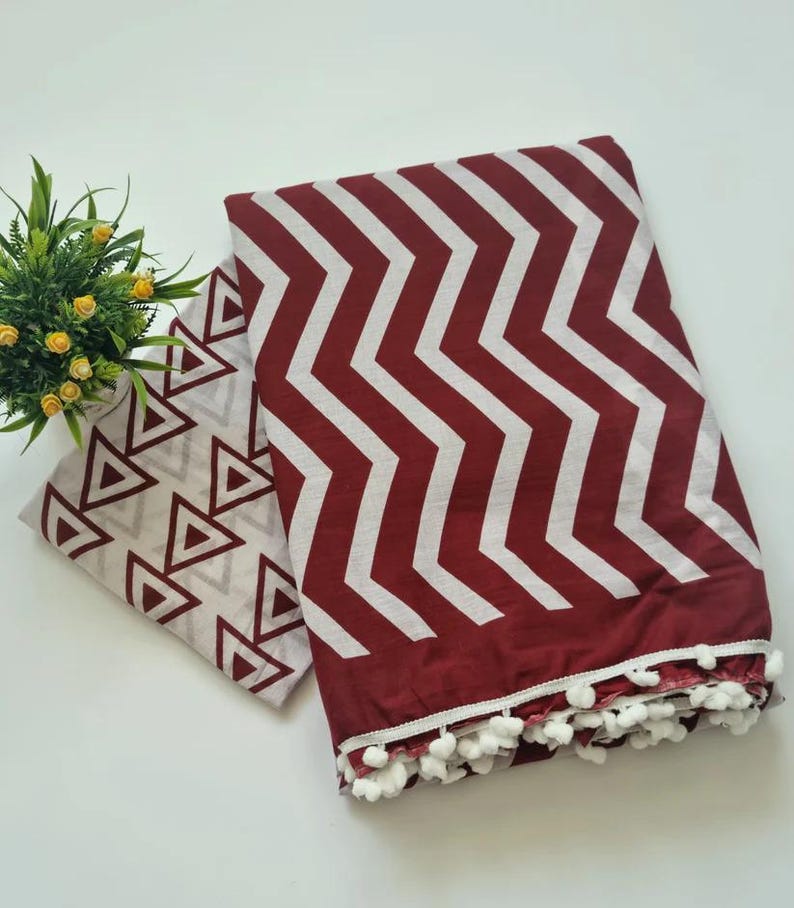 May include: A maroon and white cotton scarf with a chevron pattern and white pom-poms around the edge. The scarf is folded in half and has a white and gray triangle pattern on the inside.