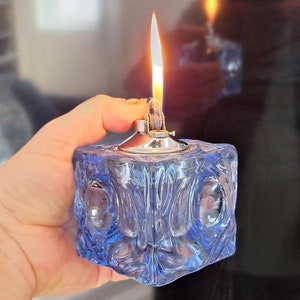 Op de afbeelding: Een blauwe glazen tafel aansteker met een zilveren bovenkant en een vlam. De aansteker wordt in een hand gehouden.