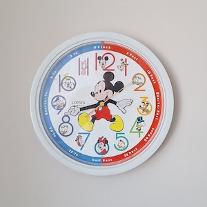May include: A white Mickey Mouse character clock with red and blue accents. The clock features Mickey Mouse walking with his arms outstretched. The numbers 1 through 12 are displayed around the clock face. The clock is on a white wall.